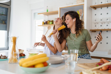 Cheerful Female Friends Having Fun Singing With Phone As Microphone During Morning Meal Preparation...