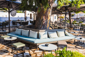 Close-up shot of a chic outdoor cafe lounge area featuring modern, comfortable seating and tables, artistically arranged around a central tree on a sunny day.