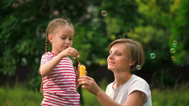 Happy mother with joyful daughter blows bubbles in park among trees on vacation. Preschooler girl waits till mother blows bubbles in meadow at weekend. Mother and child spend time blowing bubbles
