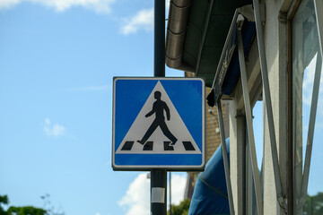 A prominent traffic sign shows a pedestrian icon on a blue background, alerting drivers to a nearby crossing. The structure is partially visible alongside the sign in bright daylight