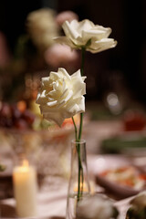 White roses in glass vases and burning candle on table with dark background. Elegant floral arrangement for wedding or celebration decor. Concept romance, luxury, event decoration.