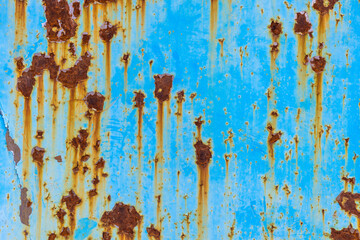 Close-up of a weathered blue metal sheet with significant rust patches and streaks running vertically. The paint is peeling and faded, revealing the underlying corrosion.