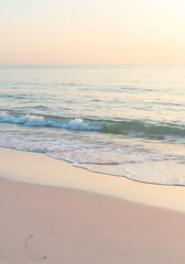 Gentle waves lap onto a sandy beach under a soft, pastel-colored sky at sunrise or sunset.