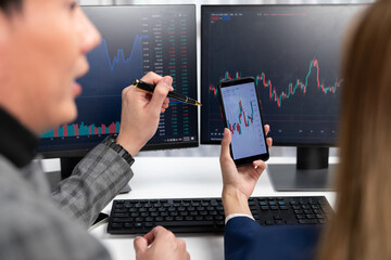 Cropped pointing hand with pen in businessman, checking with holding smartphone by woman investor online website program application with dynamic stock market at home office at back side. Infobahn.