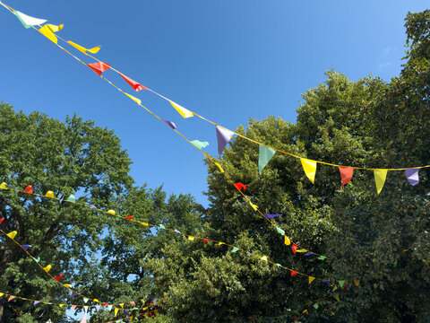 Colorful festive pennant flags hanging on strings against blue summer sky and green trees creating bright decorative background for holiday celebration and cheerful outdoor events