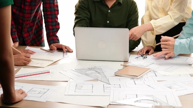 Architect and safety engineer discussion about building blueprint displaying on laptop on table with architectural document and wooden block scatter around at modern office. Closeup. Delineation.
