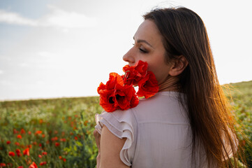 Naklejka premium Backlit woman in a light dress holding red poppies in a blooming wildflower meadow at golden hour; serene summer landscape, romantic mood, mindfulness, nature, freedom, lifestyle