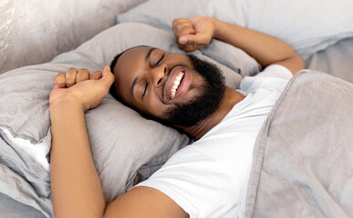 Good Morning. Satisfied young African American man stretching arms after wake up, feeling rested, happy bearded black guy sleeping lying on the gray linen flax bedsheets at home, relaxing in bedroom