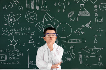 Thoughtful little boy in lab coat and protective glasses near chalkboard at chemistry lesson in classroom