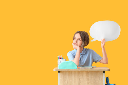 Happy schoolboy with stationery and blank speech bubble sitting on desk against yellow background