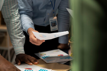 Close up of colleagues attend an after hours business meeting for analysis on files, exchange infographics and performance indicators to ensure project agenda planning. Late night briefing.