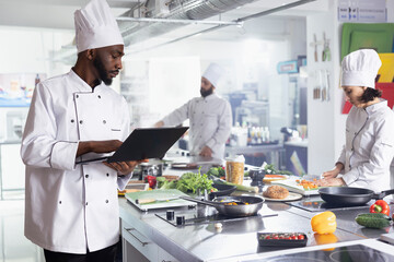 African american chef in a modern kitchen using laptop to discover new dishes and recipe tips, preparing a new fine dining menu. Young professional cook working in gastronomy, culinary arts.
