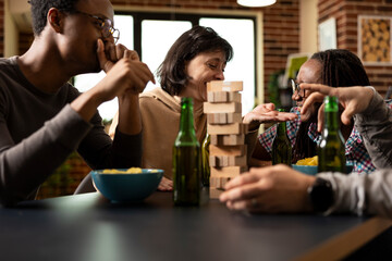 Group of multiethnic friends bonding through laughter and jokes, surrounded by snacks and beverages in cozy living space. Cheerful diverse group having fun, playing board games in brick wall room.