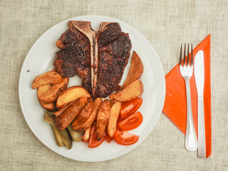 Meal with high quality T bone steak, wedges, fresh tomatoes and pickles on white plate and fork and knife on red napkin. Top version of beef strip loin with bone in for a special great flavor.