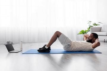 Fototapeta premium Smiling young black man with beard in white sportswear do abs exercises and watch video on laptop on floor on mat in living room interior. Fitness lesson or online conference with trainer using device