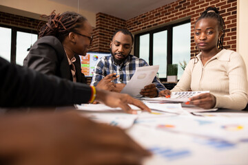 Black professionals collaborate at table, reviewing and analyzing financial charts and reports during business evaluation meeting in modern workspace. Focused team members work together in office.