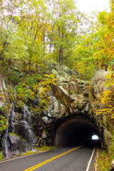A winding mountain road leads to a dark tunnel entrance, with a small waterfall cascading down the rugged rock face on the left, all framed by vibrant green and yellow autumn foliage in Shenandoah Nat