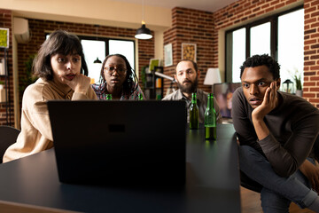 Four multicultural young adults sit closely, eyes fixed on laptop screen, reacting with astonishment during thrilling film scene. Diverse friends react to tense movie moment on device.
