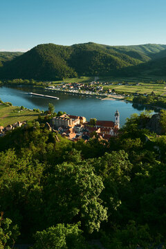 Vertical sunny view from above of a baroque church, historic town, forests, vineyards, hills, and the Danube River with a passing barge in Wachau Valley, Austria.