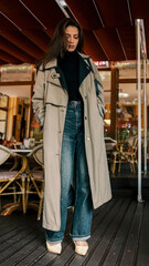 Fototapeta premium Full-length portrait of fashionable woman wearing beige trench coat, wide-leg jeans, and heels, standing thoughtfully in outdoor cafe setting