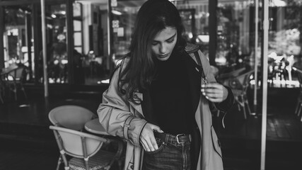 Fototapeta premium Young woman in trench coat and high-waisted jeans, standing at an outdoor café while adjusting her hair, looking down in a candid, stylish moment