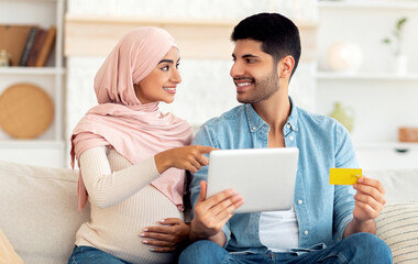 Pregnancy and online shopping concept. Muslim pregnant spouses using digital tablet and credit card purchasing goods in internet together, sitting on sofa, copy space