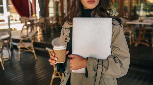Close-up of elegant woman in beige trench coat holding silver laptop and takeaway coffee in outdoor café setting