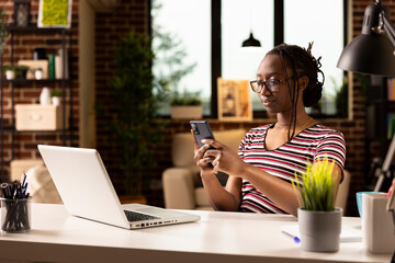 Focused african american woman with glasses multitasking from home, checking smartphone messages while working on laptop. Black female entrepreneur browsing social media and replying to texts.