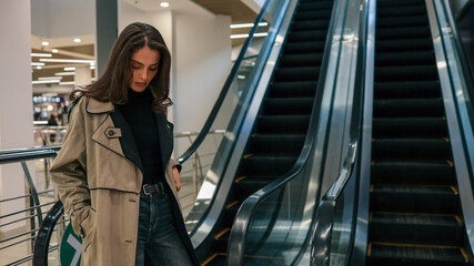 Obraz premium Young stylish woman in beige trench coat and jeans walking past an empty escalator inside a modern shopping mall, looking down thoughtfully with hands in her pockets