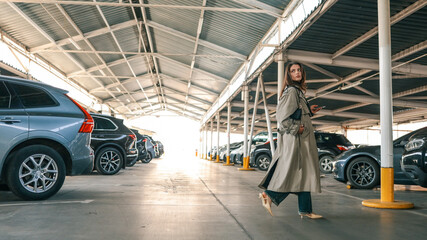 Young woman walking through a covered parking lot, wearing a long trench coat and holding a smartphone. She appears focused while surrounded by parked vehicles