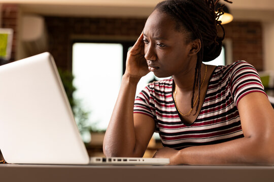 Close up of tired african american freelancer leaning forward with pained expression, touching head in frustration. Emotionally drained businesswoman struggles with work stress at home.