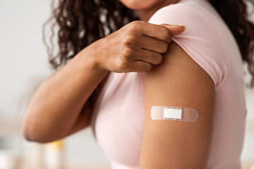 Virus protection, COVID-19 immunization. Happy young pretty african american female showing arm with band-aid after getting vaccination in modern kitchen interior, cropped, close up, free space