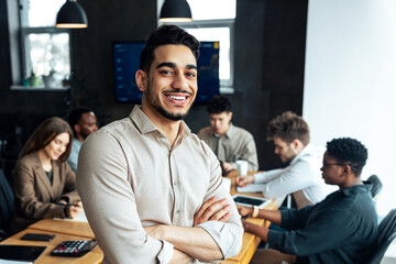 Successful Person. Portrait of confident smiling bearded businessman sitting leaning on desk in office, posing with folded arms and looking at camera, colleagues working in blurred background