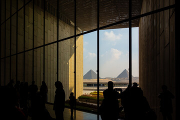 A group of people are silhouetted inside a modern building, looking out a large window at a pyramid...