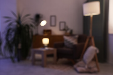 Blurred view of dark living room with glowing lamps, armchair and table in evening