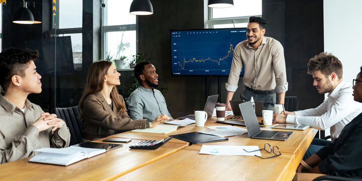 Business Presentation. Smiling Young Businessman Giving Speech During Seminar With Coworkers In Office, Standing At Desk In Boardroom, Diverse People Sitting At Table And Listening To Speaker