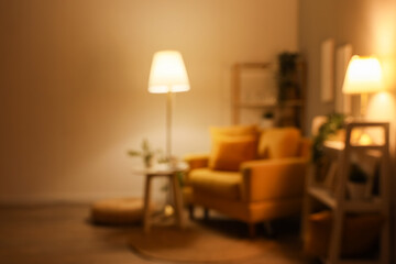 Blurred view of dark living room with glowing lamps, armchair and shelf units in evening