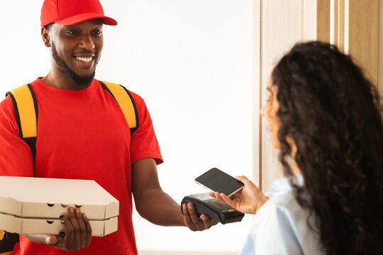Fast Food Delivery, Cashless Payment. Smiling black male courier wearing red uniform holding Point of Sale terminal and pizza boxes, female consumer paying using smartphone with blank screen
