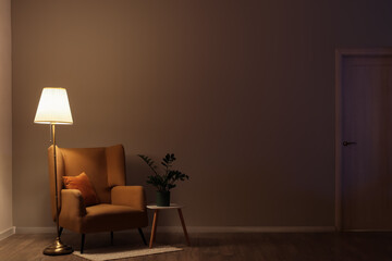 Interior of dark living room with glowing lamp, armchair and table in evening