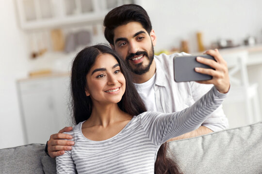 New normal, selfie, video call during social distancing. Happy attractive young arab lady and bearded guy take photo on smartphone, look at web camera, on couch in living room interior, empty space