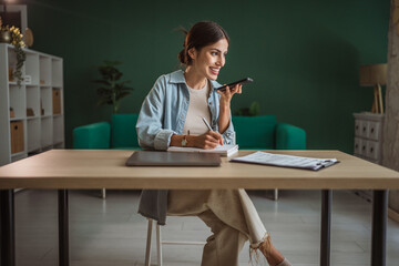 Happy entrepreneur recording voice message and taking notes at home office