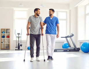 Smiling patient with crutches walks with physical therapist in bright rehabilitation gym.