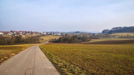 View along a rural path with fields showing early green growth, leading towards a small village in the background.