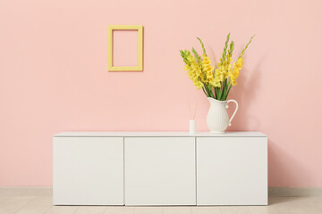 Vase with beautiful yellow gladiolus flowers on chest of drawers near pink wall