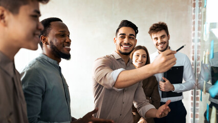 Presentation And Teamwork Concept. Creative group of smiling professional business people brainstorming, putting sticky post-it notes on glass wall in office, discussing new ideas analysing strategy