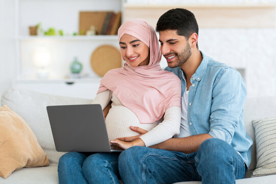 Loving pregnant muslim couple using laptop computer, watching movie together at home, resting on sofa in living room, expecting arab lady spending time with her husband, empty space - Powered by Adobe