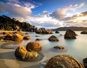 Serene coastal scene at sunrise, featuring large, smooth rocks gently resting in tranquil shallows, bathed in warm golden hues, with a backdrop of verdant hills and a vibrant sky filled with clouds.