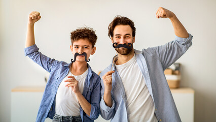 Son and strong dad showing biceps, holding fake moustache on sticks and smiling to camera. Father and boy having fun, spending time together at home.