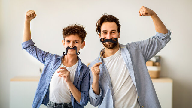 Son and strong dad showing biceps, holding fake moustache on sticks and smiling to camera. Father and boy having fun, spending time together at home.