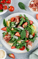Italian arugula salad with prosciutto, strawberries, melon, cheese on gray background. Top view. 